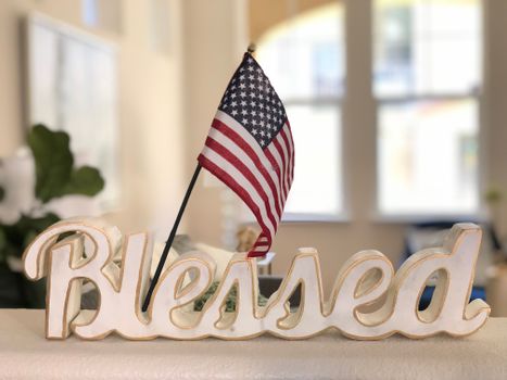 Decorative sign with American flag and 'Blessed' text on a blurred indoor background.